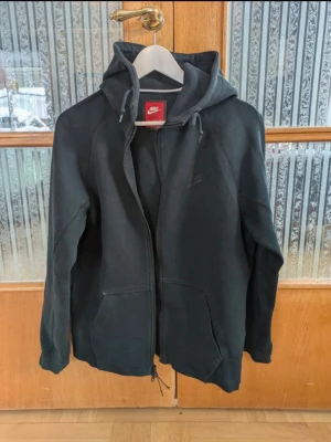 Svart hoodie med dragkedja från Nike - Svart hoodie från Nike med hel dragkedja, huva med snörning och två stora fickor framtill. Tillverkad i mjuk bomullsblandning, perfekt för en avslappnad och sportig stil. Diskret Nike-logga på bröstet och ribbade muddar vid ärmslut och nederkant.