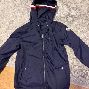 Moncler vindjacka - Snygg vindjacka från Moncler med huva. Jackan har Moncler-logga på ärmen och rödvit rand på huvan. Inga tecken på användning. Storlek S men kan passa M
