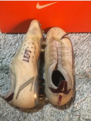 Nike Mercurial vita fotbollsskor - Säljer ett par Nike Mercurial fotbollsskor i vitt med lila detaljer och silverfärgade inslag. Skorna har en låg profil, snörning och dobbar för spel på gräs. Tillverkade i syntetmaterial för lätt vikt och bra bollkontroll. Perfekta för dig som vill ha speed på planen.