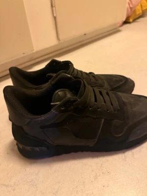 Valentino rockrunners - Snygga helsvarta sneakers med ovandel i mocka och mesh. Skorna har klassisk låg profil, svarta skosnören och en slitstark gummisula med rutmönster undertill. Perfekta för dig som gillar stilrena och diskreta skor.