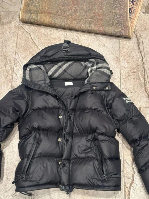 Burberry pufferjacka  - Svart Burberry jacka   Snygg och varm pufferjacka från Burberry. Bra skick, sparsamt använd.  • Svart • Skön passform • Dragkedja + knappar • Fickor med dragkedja • Klassiskt rutigt mönster i luvan  Passar till allt och är perfekt till höst/vinter.