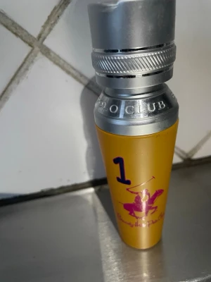 Polo Club parfym gul metallflaska - Cool parfym från Polo Club i en gul metallflaska med silverfärgat lock. Flaskan har en lila siffra 1 och en rosa polospelare på framsidan. Designen är sportig och sticker ut i badrumsskåpet.