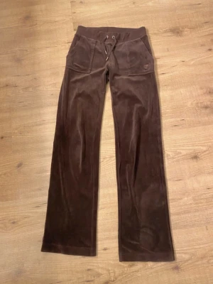 Bruna mjukisbyxor från Juicy Couture - Mörkbruna mjukisbyxor i velour från Juicy Couture med bred resårmidja och dragsko. Byxorna har två framfickor och två bakfickor med knapp. Klassisk logga på ena fickan framtill. Perfekta för en avslappnad och trendig look.