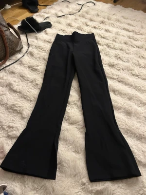 Svarta ribbade leggings med slits - Säljer ett par svarta leggings med ribbad struktur och slits nertill på benen. De har hög midja och utsvängda ben, vilket ger en snygg siluett. Perfekta att styla med sneakers eller boots för en trendig look.