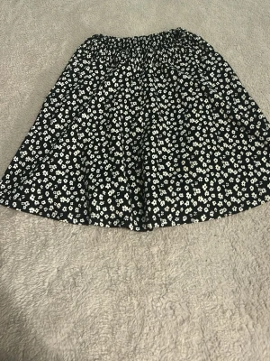 Svart blommig kortkjol från 157 - Supersöt svart kortkjol från 157 med vitt blommönster och elastisk midja. Kjolen har en luftig passform och är perfekt för dig som gillar en enkel men ändå trendig stil. Materialet känns lätt och skönt mot huden. Jag har satt resår i midjan men passar en S