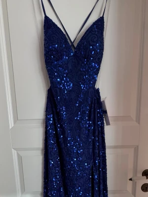 Marinblå paljettklänning med smala band - Snygg marinblå långklänning med smala axelband och v-ringning. Klänningen är täckt av glittriga paljetter och har öppen rygg med korsade band. Perfekt för dig som vill sticka ut på bal eller fest. Materialet känns lyxigt och skimrande. Storlek 34 men sitter som 36/38!
