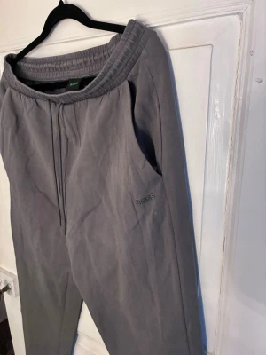 Grå mjukisbyxor från Hugo Boss - Säljer ett par grå mjukisbyxor från Hugo Boss med broderad logga på benet. Byxorna har resårmidja med dragsko och raka ben. Tillverkade i mjukt bomullsmaterial som är sköna att ha på sig. Perfekta för chill dagar eller när du vill vara bekväm.