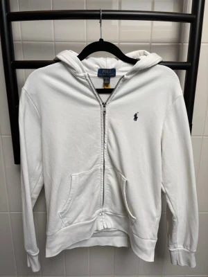 Vit hoodie från Polo Ralph Lauren - Säljer en vit hoodie från Polo Ralph Lauren med dragkedja framtill och klassisk blå logga broderad på bröstet. Hoodien har huva, kängurufickor och ribbade muddar. Tillverkad i mjuk bomull för skön känsla och stilren look.