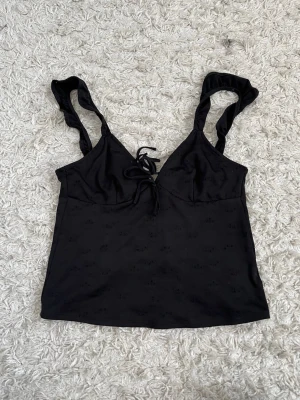 Svart linnetopp med snörning - Snygg svart linnetopp med tunna axelband och snörning både fram och bak. Toppen har en lätt v-ringad form och volangdetaljer på axelbanden. Perfekt att styla med jeans eller kjol för en trendig look.