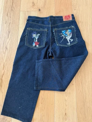 Loose fit Mörkblå jeans - Unika mörkblå jeans med raka ben och broderade motiv på bakfickorna. Jeansen har klassisk femficksdesign och kontrastsömmar. Materialet är kraftig denim och passformen är avslappnad. Perfekta för dig som vill sticka ut med coola detaljer. 