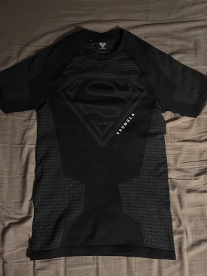 Svart Superman compression shirt  från YoungLA - Säljer en svart superman compression shirt  från YoungLA med stort Superman-logga på bröstet. Tröjan har korta ärmar, rund hals och detaljer i grått mönster på sidorna och axlarna. Tillverkad i ett stretchigt funktionsmaterial som passar perfekt till träning. Den är helt ny bara använd 