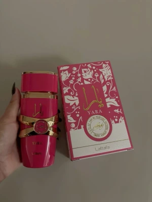 Yara parfym från Lattafa, rosa/guld - Yara parfym från Lattafa i en snygg rosa flaska med gulddetaljer och matchande ask. Flaskan har en modern, rektangulär form och dekoreras med ett guldband och emblem. Förpackningen är också rosa med vita blommönster och guldtext.