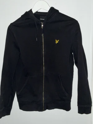 Svart zip hoodie från Lyle & Scott - Svart hoodie med dragkedja från Lyle & Scott. Klassisk design med huva och dragsko, samt kängurufickor framtill. På bröstet sitter den ikoniska gula örnloggan. Perfekt för en avslappnad och stilren look.