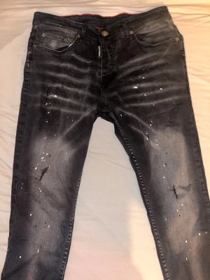 Svarta Dsquared2 jeans med splatter - Säljer ett par svarta jeans från Dsquared2 med coolt splattermönster och slitningar på benen. Jeansen har fem fickor, knappgylf och röd detalj på insidan av midjan. Materialet är klassisk denim och passformen är slim med raka ben dock jag då aldrig använt byxorna. Skriv gärna om du har fler frågor mvh.