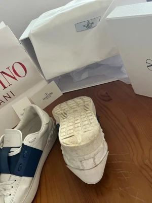 Valentino vita sneakers med blå detalj - Snygga vita sneakers från Valentino med bred blå läderdetalj över sidan. Klassisk låg modell med rund tå och vita snören. Märkeslogga på plösen och insidan. Kommer med originalkartong och dustbag.