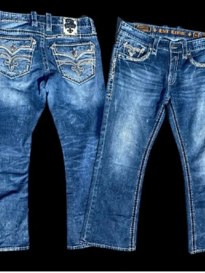 Rock Revival blå jeans med brodyr - Snygga blå jeans från Rock Revival med unika broderade detaljer på bakfickorna och kontrastsömmar. Jeansen har en klassisk femficksmodell, raka ben och slitningar för en cool look. Materialet är denim i bomull med stretch. Om fu vill ha mer info bara skriva 