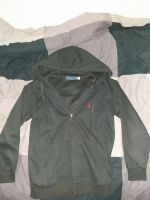 Svart hoodie från Polo Ralph Lauren - Klassisk svart hoodie från Polo Ralph Lauren med dragkedja framtill och huva med snören. Ikoniska röda loggan broderad på bröstet. Tillverkad i mjukt bomullsmaterial som är skönt att bära. På riktigt använd 2 gånger säljer för den är förliten för mig nu. (Kan finnas lite hund hår på tröjan)