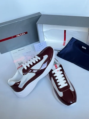 Prada sneakers vinröd och vit - Snygga sneakers från Prada i vinrött och vitt med glansiga paneler och meshdetaljer. Klassisk låg modell med vita skosnören och tjock vit sula. Märkeslogga på plösen och insidan. Perfekta för dig som vill ha en exklusiv och trendig look.