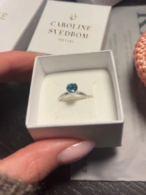 Silverring med blå sten Caroline Svedbom - Hej!🌸Snygg silverfärgad ring från Caroline Svedbom med en rund, gnistrande blå sten i mitten och små klara stenar längs bandet. Ringen har en elegant och modern design som verkligen fångar ögat. Perfekt accessoar för att lyfta din stil. Helt oanvänd! Nypris 395kr🫶
