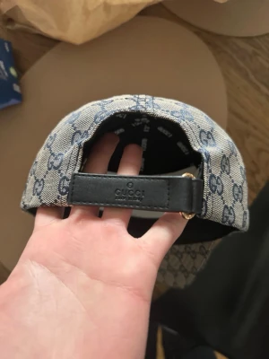  keps med GG-mönster - Snygg keps  i beige tyg med blått GG-monogram över hela kepsen. Justerbar rem bak i svart läder och klassisk -rand i rött och blåt på sidan. Perfekt för dig som vill ha en exklusiv och trendig accessoar.