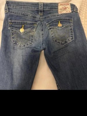 Blå True Religion jeans med kontrastsömmar - Säljer ett par blå jeans från True Religion med ikoniska kontrastsömmar och detaljerade bakfickor med lock och metallknappar. Klassisk femficksmodell i stretchigt denimtyg. Perfekta för dig som gillar streetwear och vill sticka ut med snygga detaljer.
