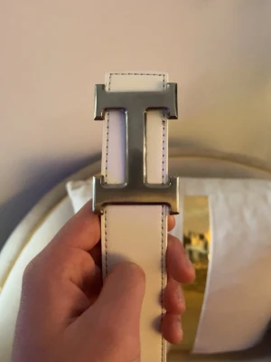 Vitt H’e’r’m’e”s skärp med silverfärgat H spänne - Snyggt vitt skärp från H’e’r’m’e”s med ett silverfärgat H format spänne. Bältet har stilrena svarta sömmar längs kanterna. Perfekt accessoar för att lyfta din outfit.