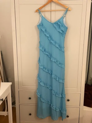 Ljusblå långklänning med volanger - Säljer en ljusblå långklänning med smala axelband och diagonala volangdetaljer över hela klänningen. Klänningen har en mjuk och flytande form som ger en romantisk känsla. Perfekt för sommarens alla tillfällen!