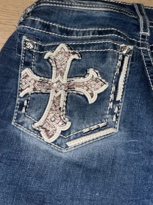 Blå jeans med broderade kors - Säljer ett par blå jeans från Miss Me med unika broderade kors i vitt och rosa på bakfickorna. Jeansen har kontrastsömmar och detaljerade fickor med bling. Materialet är klassisk denim i bomull med stretch för extra komfort.