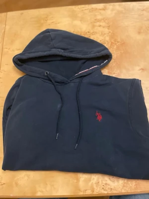 Mörkblå hoodie från Polo  - Snygg mörkblå hoodie från Polo  med klassisk röd broderad logga på bröstet. Hoodien har dragsko i huvan och är tillverkad i mjukt bomullsmaterial. Perfekt för en avslappnad och stilren look.
