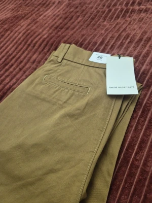 These Glory Days T Aimon straight olivgröna byxor, storlek 48 - Snygga olivgröna chinos från These Glory Days i klassisk rak modell. Byxorna har bälteshällor, diskreta bakfickor och är tillverkade i ett mjukt bomullstyg. Perfekta för en clean och stilren look.