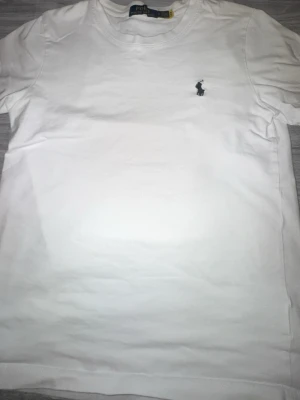 Vit t-shirt från Polo Ralph Lauren - Klassisk vit t-shirt från Polo Ralph Lauren med den ikoniska svarta polospelaren broderad på bröstet. T-shirten har rund hals och korta ärmar, tillverkad i mjuk bomull för en skön och stilren look.