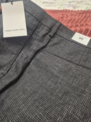 These Glory Days Kelly Structure Cuff gråa byxor, storlek 46 - Snygga grå kostymbyxor från These Glory Days med diskret rutmönster. Byxorna har raka ben, bälteshällor och klassisk knappstängning. Perfekta för en stilren look. Materialet känns mjukt och har en elegant struktur.