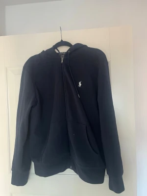 Marinblå hoodie från Polo Ralph Lauren - Snygg marinblå hoodie från Polo Ralph Lauren med dragkedja framtill och klassisk vit logga på bröstet. Hoodien har huva med dragsko och två stora fickor framtill. Tillverkad i mjukt bomullsmaterial som är skönt att bära.