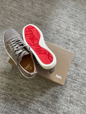 Gråa Louboutin sneakers i mocka - Snygga mock distincta sneakers från Christian Louboutin i grått med klassisk röd sula och vita kanter. Skorna har rund tå, gråa skosnören och diskret branding på insidan. Perfekta för dig som vill ha en clean och exklusiv look.