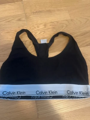 Svart sport-bh från Calvin Klein - Säljer en svart sport-bh från Calvin Klein med bred vit resår och logga runt om. Bh:n har brottarrygg och är tillverkad i mjukt bomullsmaterial för extra komfort. Perfekt för en avslappnad och sportig stil.