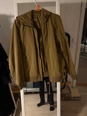 Olivgrön vindjacka med huva - Snygg olivgrön vindjacka med huva och dragkedja framtill. Jackan har två sidofickor och ribbade muddar vid ärmslut och nederkant. Perfekt för blåsiga dagar och enkel att matcha med olika outfits.