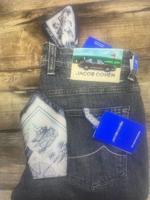 Jacob Cohen svarta jeans med detaljer - Snygga svarta jeans från Jacob Cohen med klassisk femficksmodell och silverfärgade knappar. Jeansen har en unik patch med bilmotiv bak och broderad J på myntfickan. Tillverkade i mjuk denim och levereras med en exklusiv scarf i fickan.