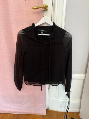 Svart blus  - Snygg svart blus från Vero Moda med transparenta långärmade ärmar och volangdetaljer vid halsringningen. Blusen har knytband fram och är gjord i ett lätt, tunt material som ger en luftig känsla. 