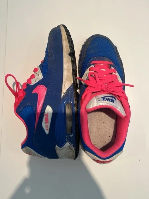 Nike Air Max blå/rosa sneakers - Nike Air Max sneakers i blått med rosa detaljer och vita inslag. Skorna har en klassisk låg siluett, mesh och mockaliknande material på ovandelen samt synlig Air Max-dämpning i sulan. Rosa snören och innersula ger en färgstark look. Priset är inte hugget i sten 
