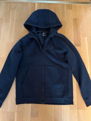 Svart Nike hoodie med dragkedja - Säljer en svart hoodie från Nike med dragkedja framtill och två stora fickor. Tröjan har huva och diskret Nike-logga på bröstet. Baksidan har ett quiltat parti i ett glansigare material som ger en sportig look. Perfekt för dig som gillar streetwear och enkel stil.