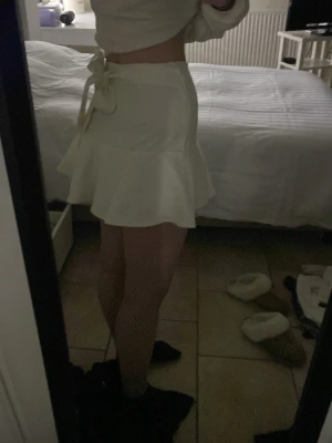 Vit kortkjol med volangkant - Säljer en vit kortkjol med volangkant med en snygg rosettdetalj i midjan som man kan välja själv om man vill ha den fram eller bak. Kjolen är helt oanvänd och har shorts under. Kjolen har en dragkedja