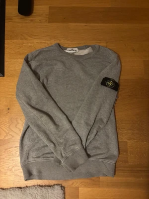 Grå sweatshirt från Stone Island - Säljer en grå sweatshirt från Stone Island med klassisk rund hals och ribbade muddar. Tröjan har den ikoniska svarta patchen med logga på vänster ärm. Perfekt för en avslappnad streetwear-look. Den har lite smådefekter vid mudden som man kan se på bilderna. Annars är den i fint skick.
