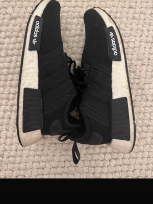 Svarta Adidas NMD_R1 sneakers - Säljer ett par svarta Adidas NMD_R1 sneakers med vit sula och svarta detaljer på sidan. Skorna har stickad ovandel i mesh, klassiska tre ränder och snörning. Märket syns tydligt på plösen och hälen. Sparsamt använda. 
