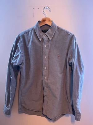 Grå skjorta från Polo Ralph Lauren - Klassisk grå skjorta från Polo Ralph Lauren med button-down krage och broderad logga på bröstet. Skjortan har långa ärmar och vita knappar framtill. Perfekt till en clean och stilren look.