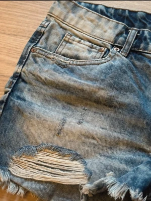Blå slitna jeansshorts med fransar - Säljer ett par blå jeansshorts med slitna detaljer och fransig kant. Shortsen har klassisk femficksdesign, knappgylf och en cool, avslappnad look. Perfekta för dig som gillar en edgy stil.