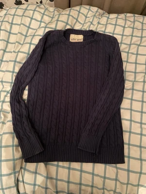 Mörkblå kabelstickad tröja Iconic Sport - Säljer en mörkblå kabelstickad tröja från Iconic Sport. Tröjan har rund halsringning, långa ärmar och ribbade muddar vid ärmslut och nederkant. Perfekt för dig som gillar klassisk och enkel stil.