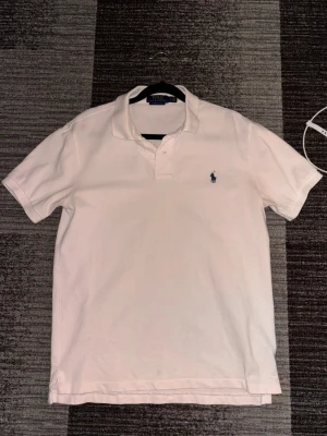 Vit pikétröja från Ralph Lauren - Klassisk vit pikétröja från Ralph Lauren med blå broderad logga på bröstet. Tröjan har krage, korta ärmar och knappar vid halsen. Passformen är slim och materialet känns som bomull. Perfekt till en clean och stilren look.