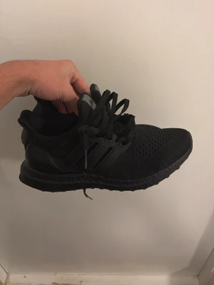 Svarta Adidas Ultraboost sneakers - Säljer ett par helsvarta Adidas Ultraboost sneakers med stickad ovandel och klassiska tre ränder på sidan. Skorna har en bekväm och responsiv Boost-sula samt snörning för perfekt passform. Perfekta för dig som gillar stilrena och sportiga sneakers.