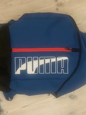 Blå ryggsäck från Puma - Snygg blå ryggsäck från Puma med stor vit logga på framsidan och en röd dragkedja. Väskan har vadderade axelband i blått och grått för extra komfort. Perfekt för skola eller träning och har en sportig look.