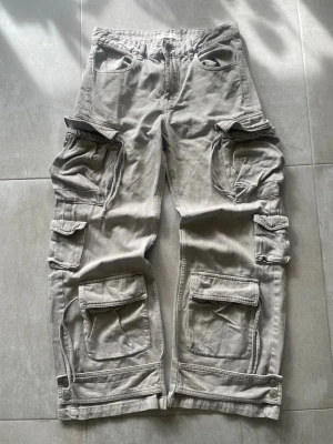 Grå cargobyxor med stora fickor pull & bear - Säljer ett par grå cargobyxor med flera stora fickor, dragkedjor och knappar vid bensluten. Byxorna har en loose fit och är tillverkade i mjuk bomull. Perfekta för dig som gillar utility-stil och vill ha plats för allt i fickorna.   Mått 📏 Totallängd: 97cm Midjelängd: 38cm Innersömm: 74cm Lårviddlängd: 26cm Skador 🔧: Inga  Storlek 🧍: EU 36  Passform: Baggy  Jag kan förhandla mig till lägre pris.🙋💵 Om du har några frågor, vänligen skriv till mig 🤗